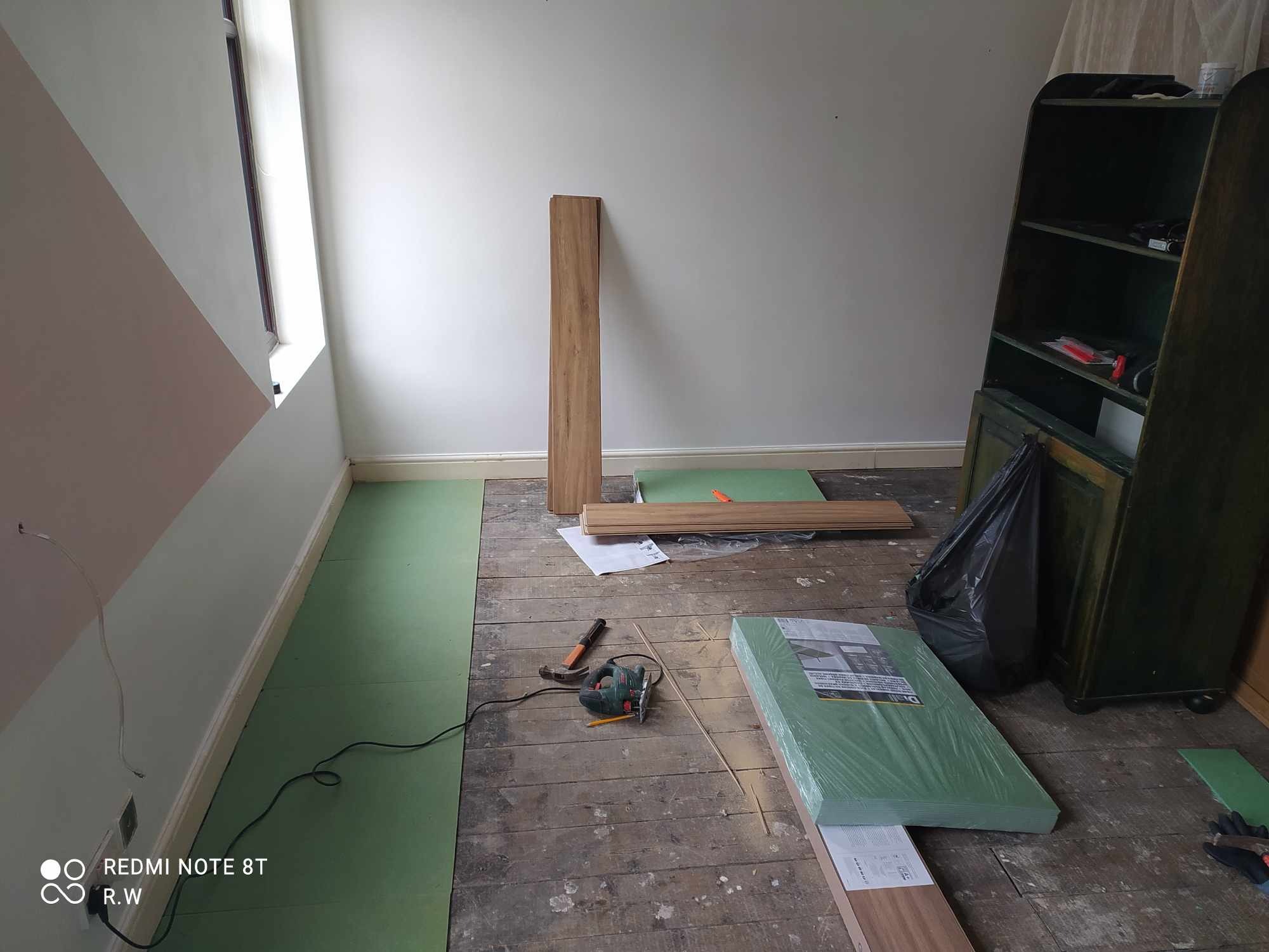 Przygotowanie do ułożenia paneli podłogowych: rozłożona zielona pianka izolacyjna, paczka paneli w folii, wyrzynarka elektryczna i młotek na starej drewnianej podłodze w pokoju z pomalowaną ścianą...