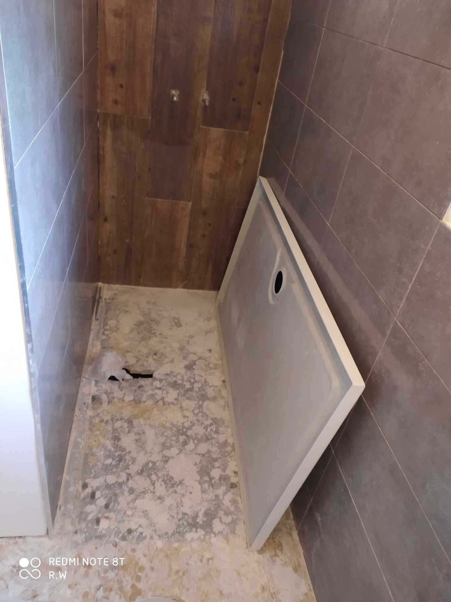 Przygotowanie do montażu brodzika w łazience: widoczny surowy beton na podłodze, częściowo położone szare płytki ścienne i drewnopodobne panele na ścianie, biały brodzik z otworem odpływowym...