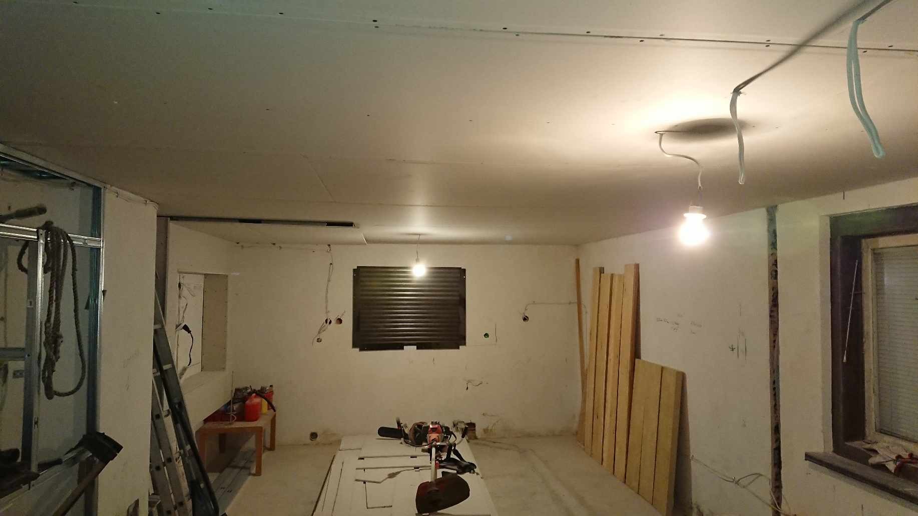 Wnętrze w trakcie prac wykończeniowych: ściany i sufit pokryte płytami kartonowo-gipsowymi, widoczne profile metalowe, narzędzia i materiały budowlane, surowa żarówka zwisająca z sufitu.