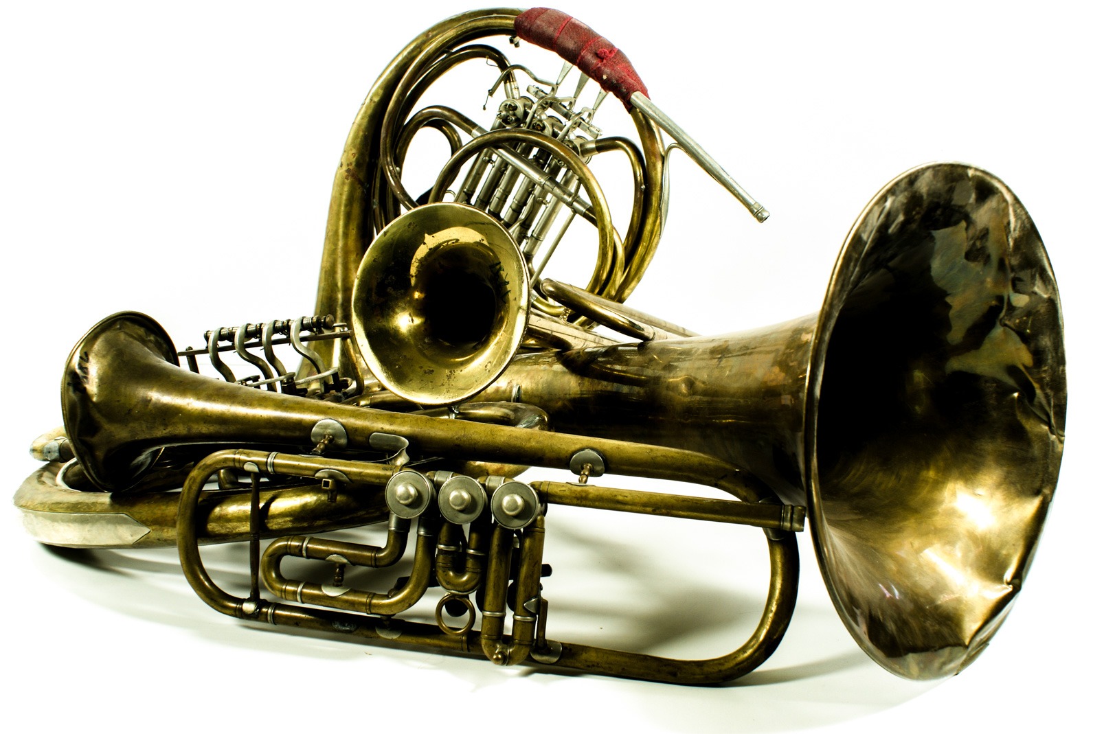 Kolekcja używanych, mosiężnych instrumentów dętych blaszanych w różnym stanie, w tym waltornia z czerwonym ochraniaczem na ustnik i tuba z widocznymi wgnieceniami na czarze, na białym tle.
