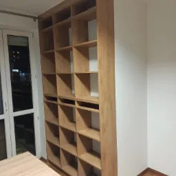 Wysoka, drewniana biblioteczka z licznymi półkami, dopasowana do wysokości pomieszczenia, stoi przy białej ścianie obok okna z widokiem na nocne miasto.