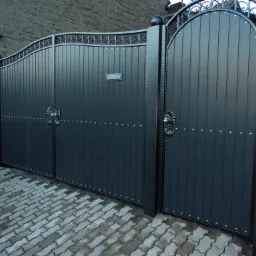 Nowoczesna, dwuskrzydłowa brama wjazdowa i furtka w kolorze antracytowym z dekoracyjnymi elementami kutymi, zamontowana na posesji z brukowanym podjazdem.