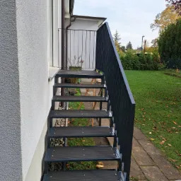 Zewnętrzne schody metalowe z ciemnoszarą balustradą prowadzące z balkonu do ogrodu, widok z dołu, w tle trawnik z opadłymi liśćmi.