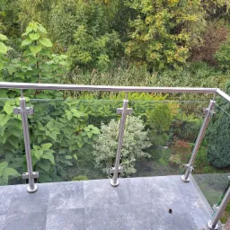 Balkon z nowoczesną balustradą ze stali nierdzewnej i szkła, widok na bujny ogród z różnorodną roślinnością, szare płytki na podłodze balkonu.