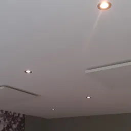 Biały sufit z zamontowanymi panelami i kilkoma punktowymi lampami halogenowymi, widoczny fragment ściany z dekoracyjną tapetą.