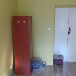 Usługi Remontowe TOMALA - Pomalowane na żółto ściany pokoju z brązową szafą z wieszakami, podłogą z paneli, białymi drzwiami i zapakowanymi materiałami budowlanymi w folii.