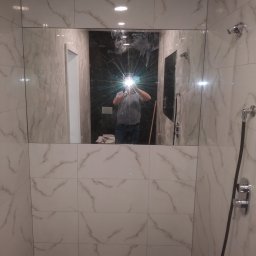 Usługi Remontowe TOMALA - Wykończona kabina prysznicowa z białymi płytkami imitującymi marmur, czarną podłogą i widocznym odbiciem osoby robiącej zdjęcie w lustrze.