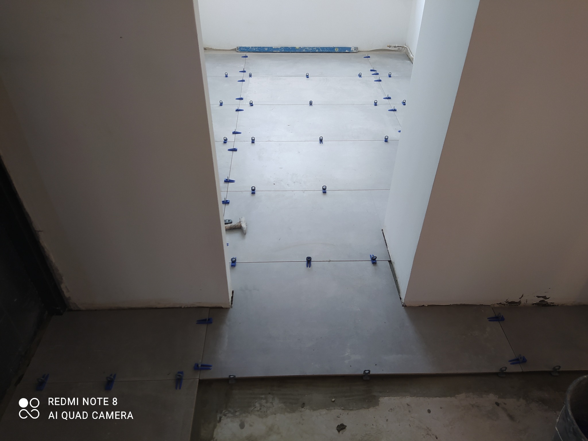 Układanie szarych płytek podłogowych w korytarzu, widoczne niebieskie klipsy poziomujące, poziomica na ścianie w tle.