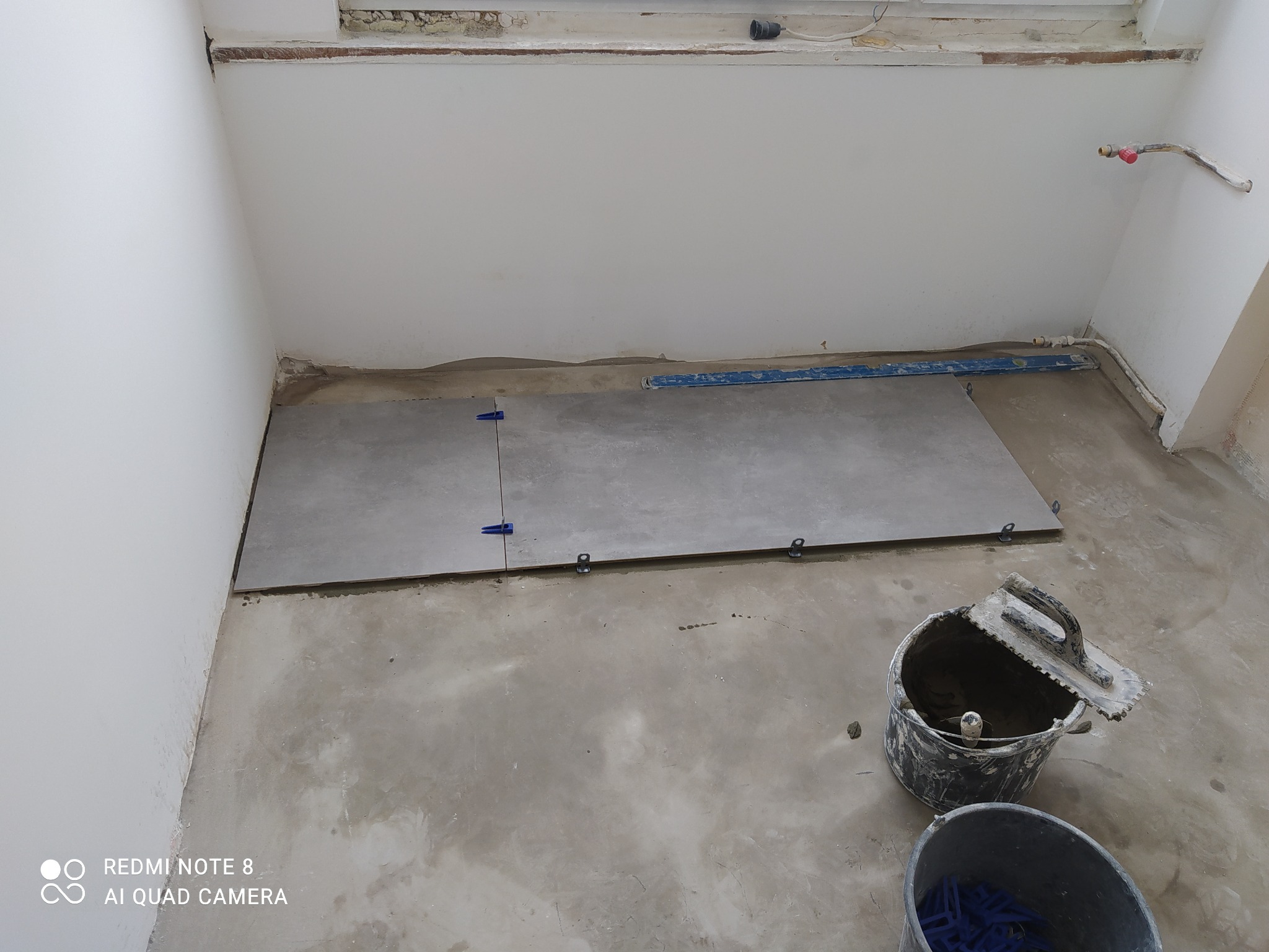 Układanie szarych płytek wielkoformatowych na podłodze w trakcie prac w Łodzi, widoczne poziomica, wiadro z zaprawą i niebieskie kliny dystansowe.