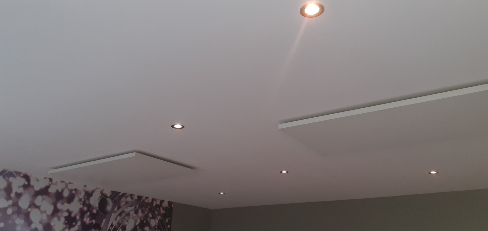 Biały sufit z zamontowanymi panelami i kilkoma punktowymi lampami halogenowymi, widoczny fragment ściany z dekoracyjną tapetą.