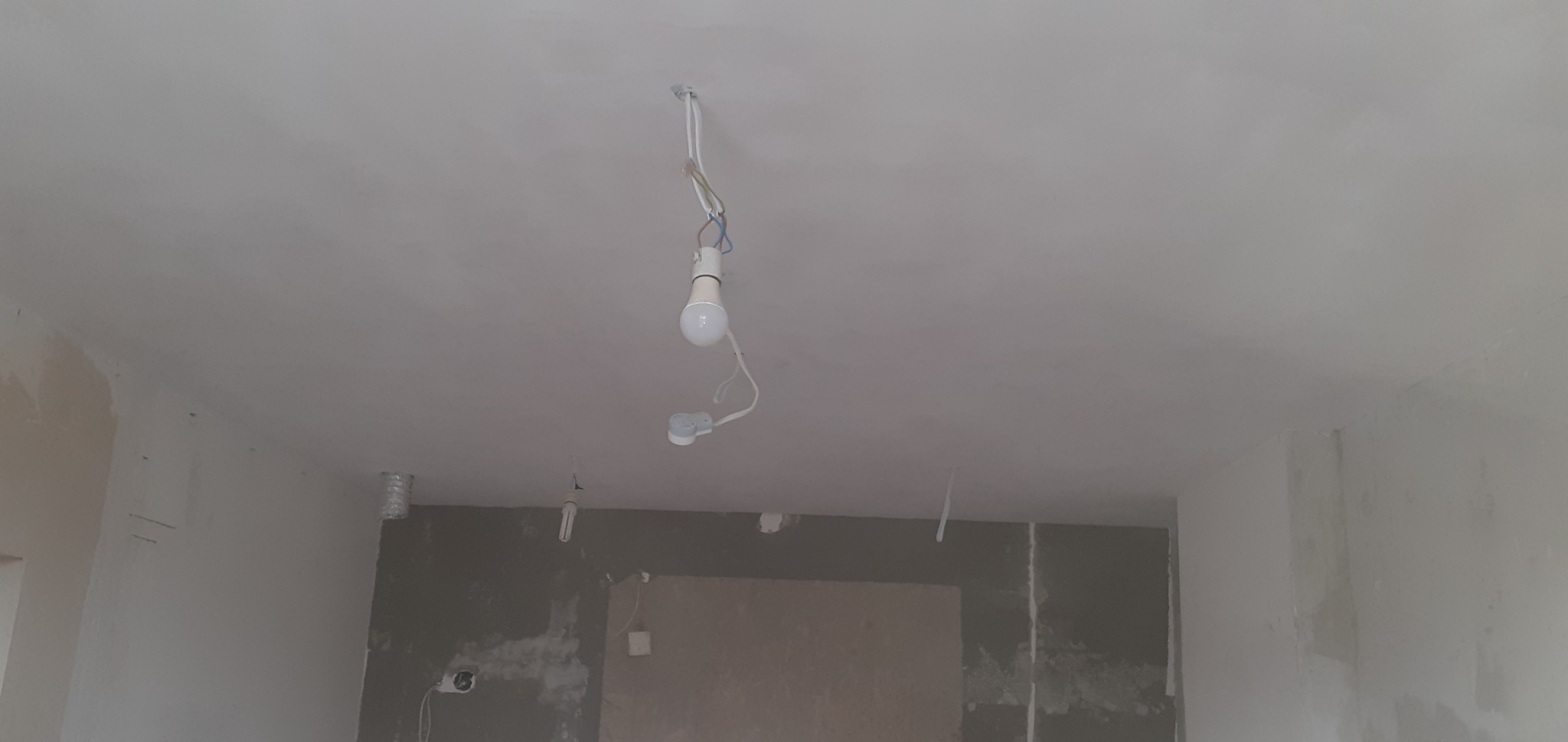 Wnętrze w trakcie prac wykończeniowych: surowy sufit i ściany po szpachlowaniu, wiszące żarówki na kablach, widoczne ślady po instalacjach.