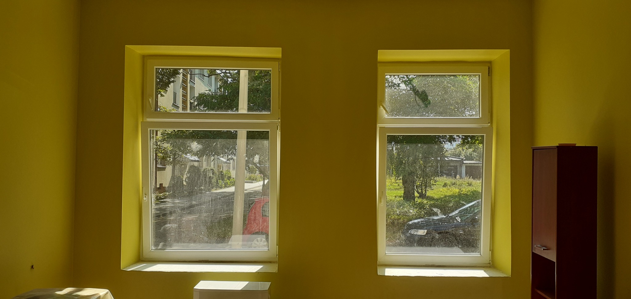 Pokój z żółtymi ścianami i dwoma oknami z widokiem na zewnątrz, w prawym rogu stoi ciemnobrązowa szafka.