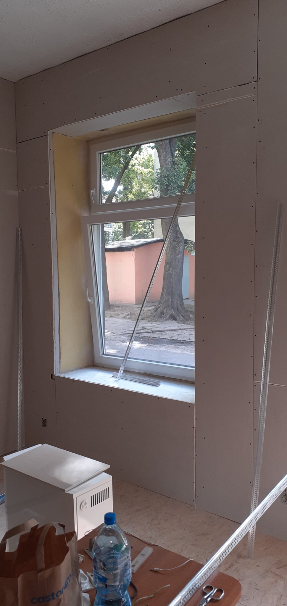 Pokój w trakcie montażu płyt kartonowo-gipsowych wokół okna, widoczne profile metalowe, butelka wody i torba budowlana na stole.