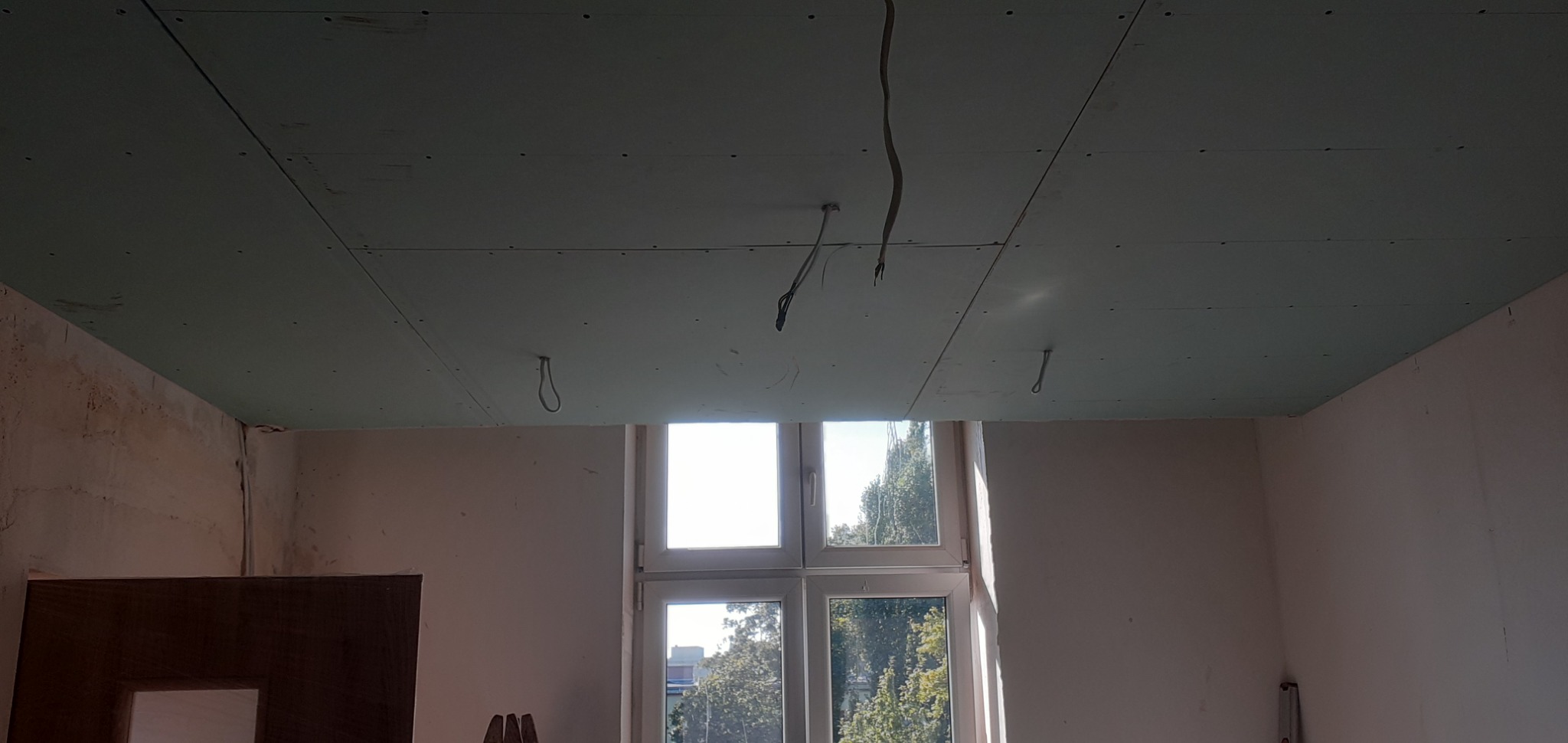 Montaż sufitu podwieszanego z płyty karton gips w pokoju z oknem, widoczne wystające przewody elektryczne i niedokończone ściany.