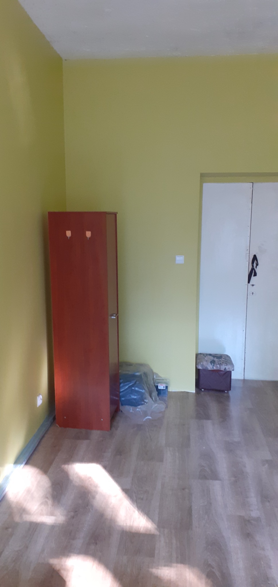 Pomalowane na żółto ściany pokoju z brązową szafą z wieszakami, podłogą z paneli, białymi drzwiami i zapakowanymi materiałami budowlanymi w folii.