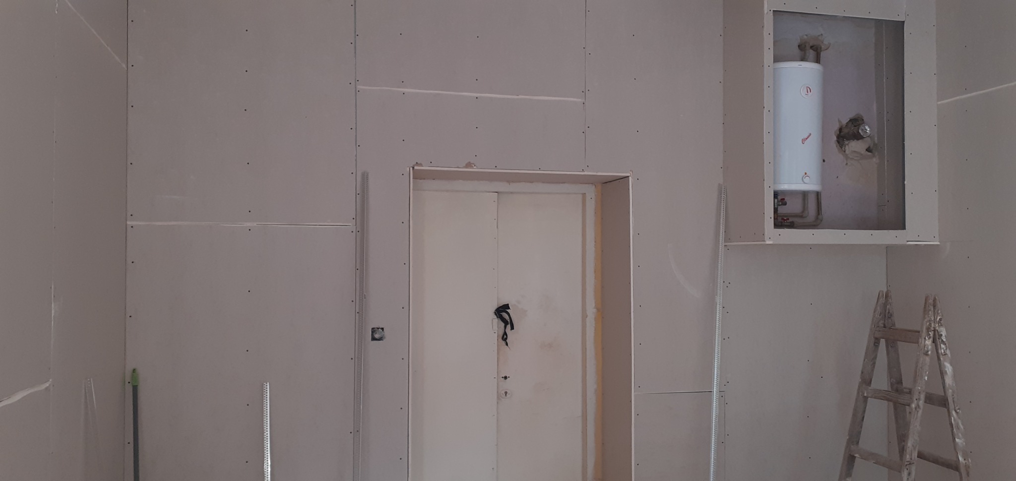 Ściana z płyt kartonowo-gipsowych w trakcie montażu, widoczne łączenia, wnęka z zamontowanym bojlerem, drabina drewniana oparta o ścianę.