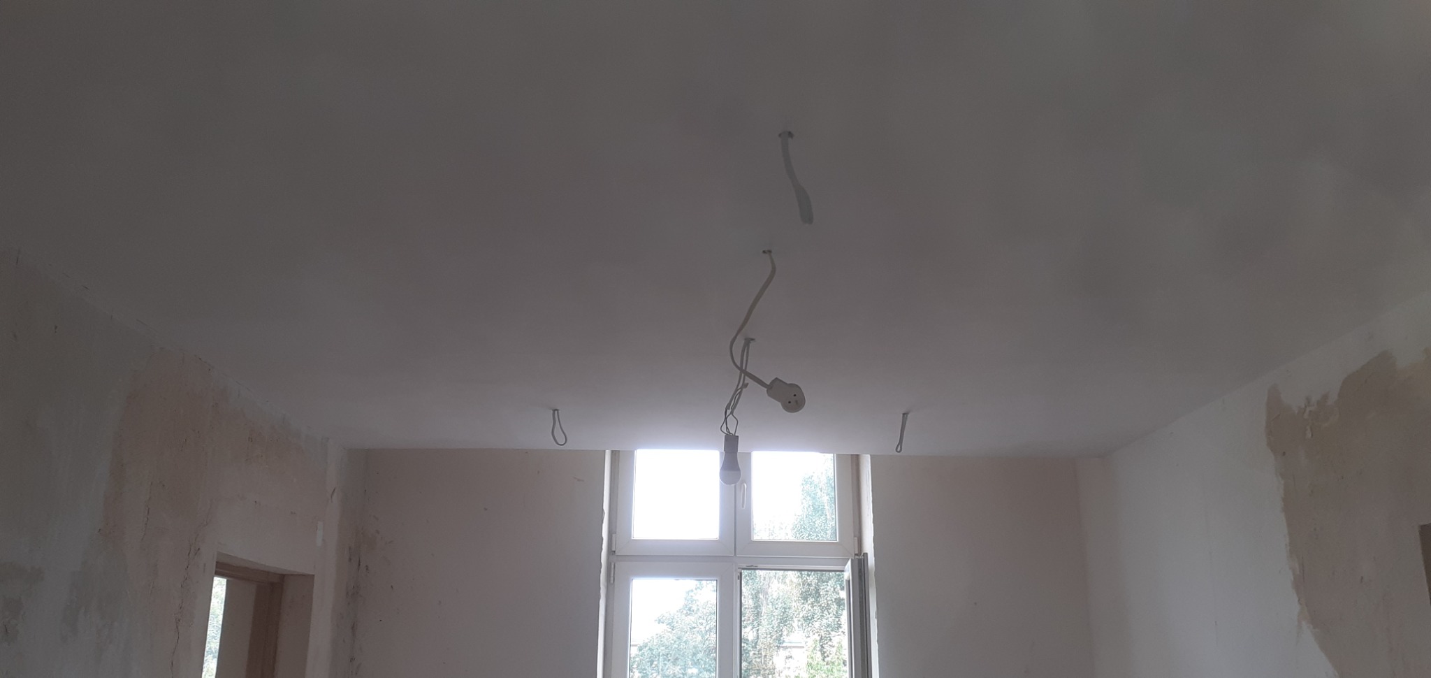 Pokój w trakcie przygotowania do malowania: ściany częściowo wyszpachlowane, sufit pomalowany na biało, widoczne wiszące kable i żarówka, okno z widokiem na zieleń.