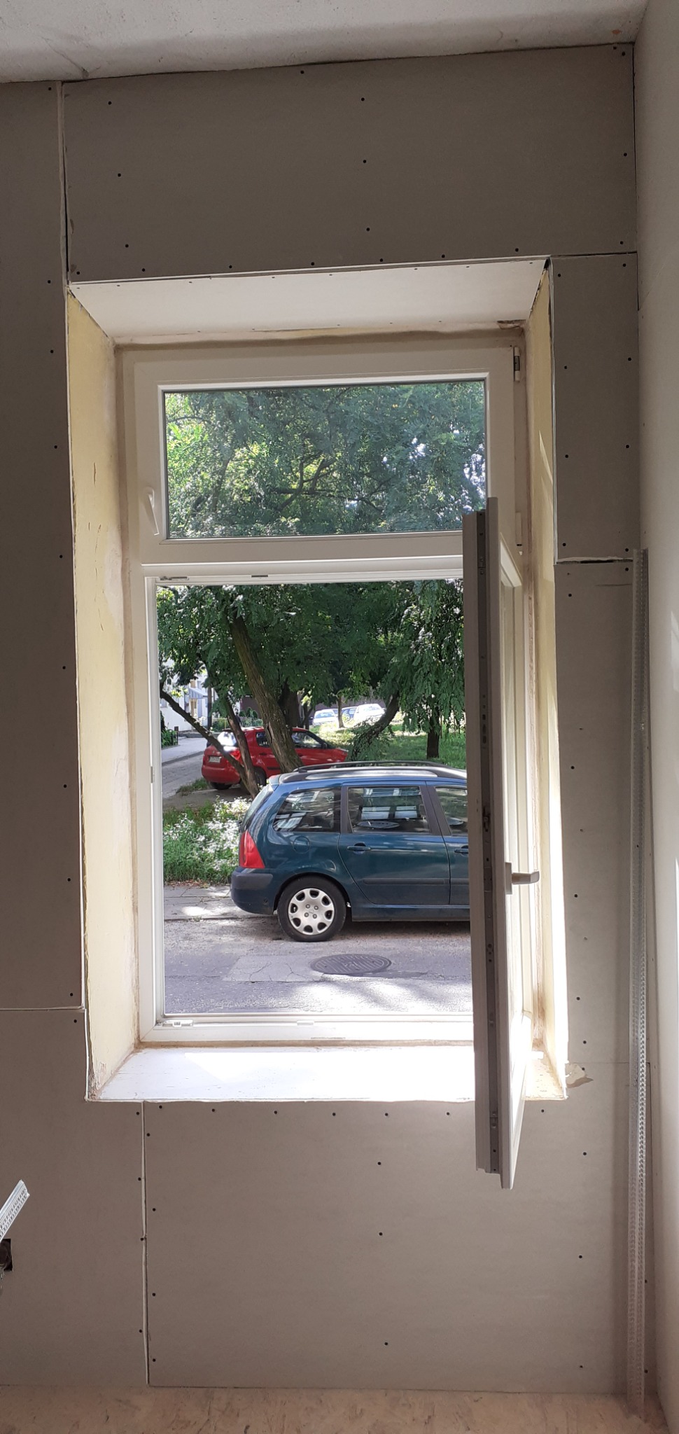 Wnętrze pokoju z oknem w trakcie montażu płyt kartonowo-gipsowych, widoczne łączenia płyt oraz częściowo otwarte okno z widokiem na ulicę i zaparkowane samochody.