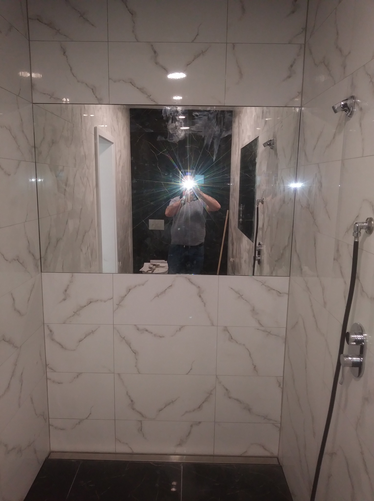 Wykończona kabina prysznicowa z białymi płytkami imitującymi marmur, czarną podłogą i widocznym odbiciem osoby robiącej zdjęcie w lustrze.