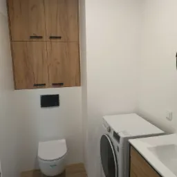 Nowoczesna łazienka z wiszącą szafką z drewnianymi frontami, podwieszaną toaletą i pralką obok umywalki z szafką w podobnym stylu, płytki podłogowe imitujące drewno, białe ściany.