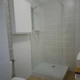 Nowoczesna łazienka z kabiną prysznicową typu walk-in, białymi płytkami z delikatnym wzorem i drewnopodobną podłogą, widoczna wisząca szafka i toaleta.