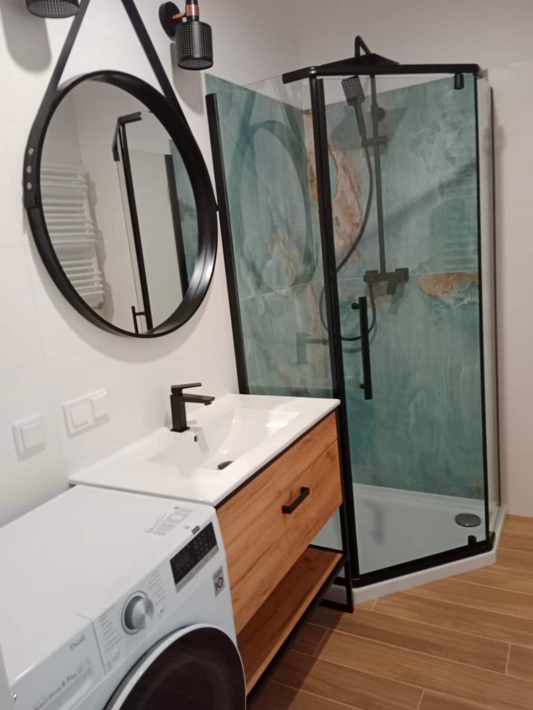 Nowoczesna łazienka po remoncie: kabina prysznicowa z zielonymi płytkami imitującymi marmur, okrągłe lustro w czarnej ramie zawieszone na pasku, umywalka z szafką z drewnopodobną okleiną i pralka.