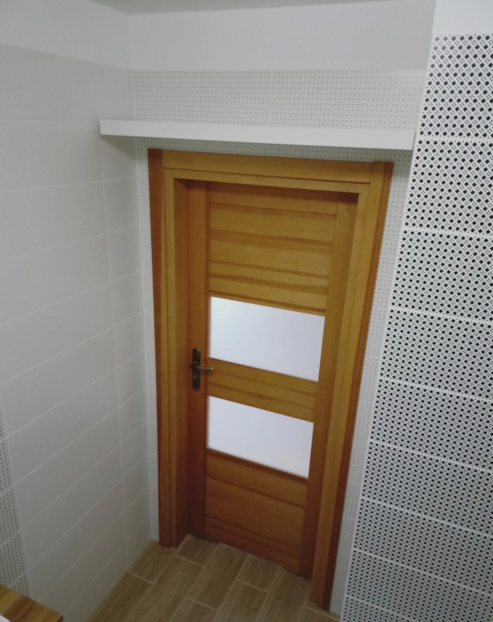 Drewniane drzwi wewnętrzne z dwoma białymi szybami, zamontowane w łazience z białymi płytkami i dekoracyjną ścianą w czarno-białe wzory.