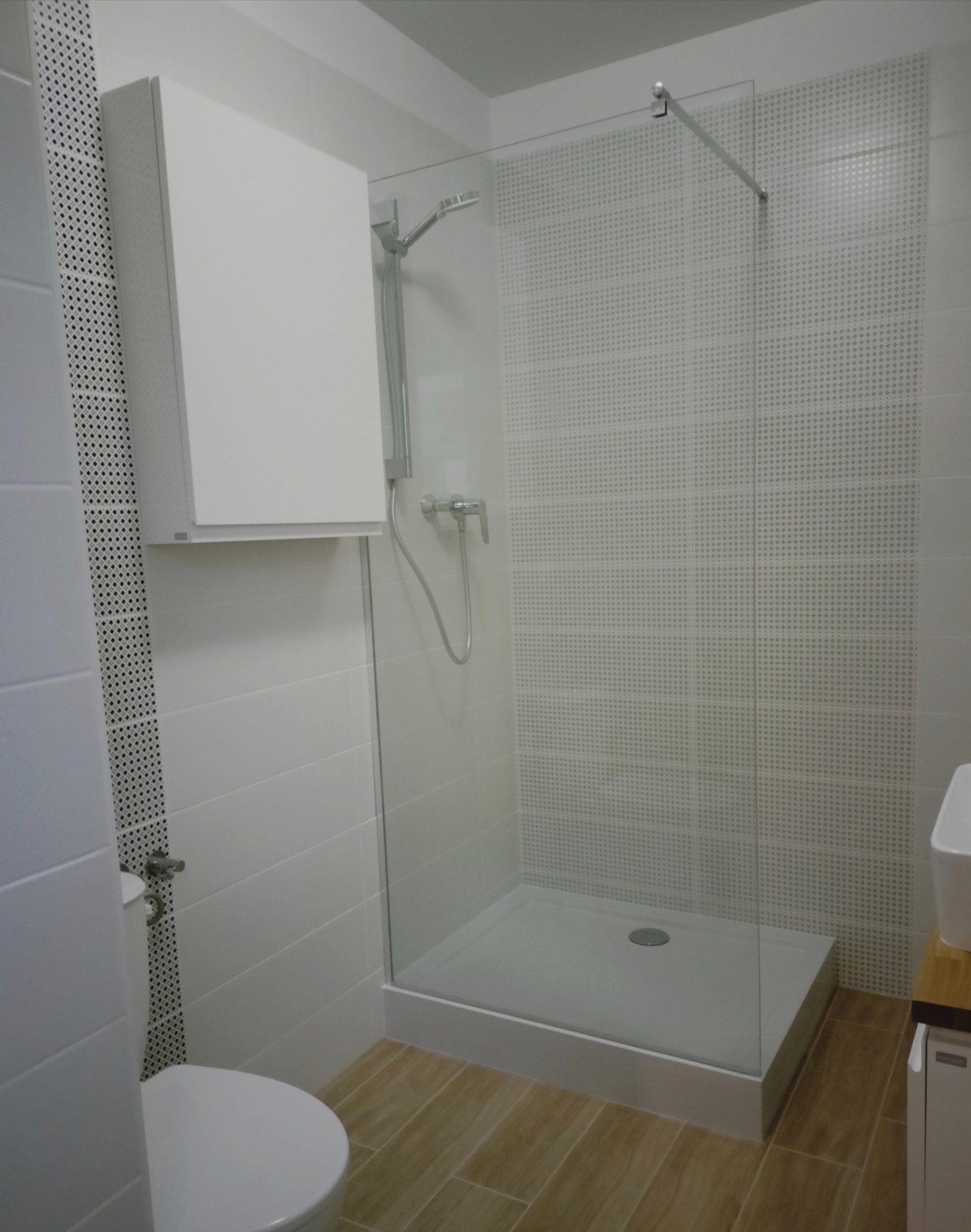 Nowoczesna łazienka z kabiną prysznicową typu walk-in, białymi płytkami z delikatnym wzorem i drewnopodobną podłogą, widoczna wisząca szafka i toaleta.