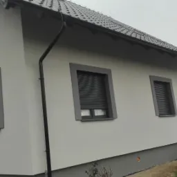 Elewacja domu jednorodzinnego w jasnych odcieniach szarości, z ciemnoszarą stolarką okienną i roletami zewnętrznymi, widoczny fragment dachu z ciemnej dachówki ceramicznej i rynna spustowa, w tle...