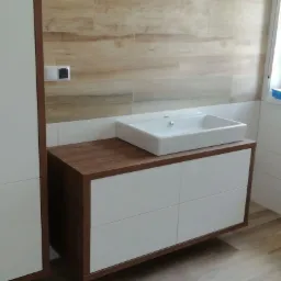 Nowoczesna łazienka z umywalką nablatową na szafce z białymi frontami i drewnianym blatem, drewnopodobne płytki na ścianie i podłodze, okno z akcesoriami malarskimi na parapecie.