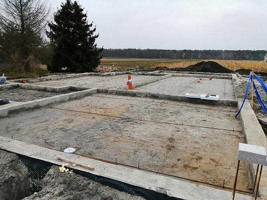Wylane betonowe fundamenty domu jednorodzinnego z wystającymi prętami zbrojeniowymi, widoczne pomarańczowe rury instalacyjne i styropian, w tle pole i las.