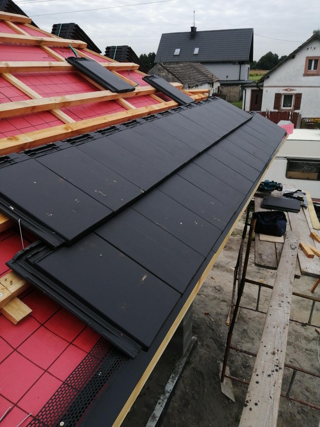 Układanie czarnej dachówki na czerwonej membranie, widoczne narzędzia i rusztowanie, w tle zabudowa jednorodzinna.