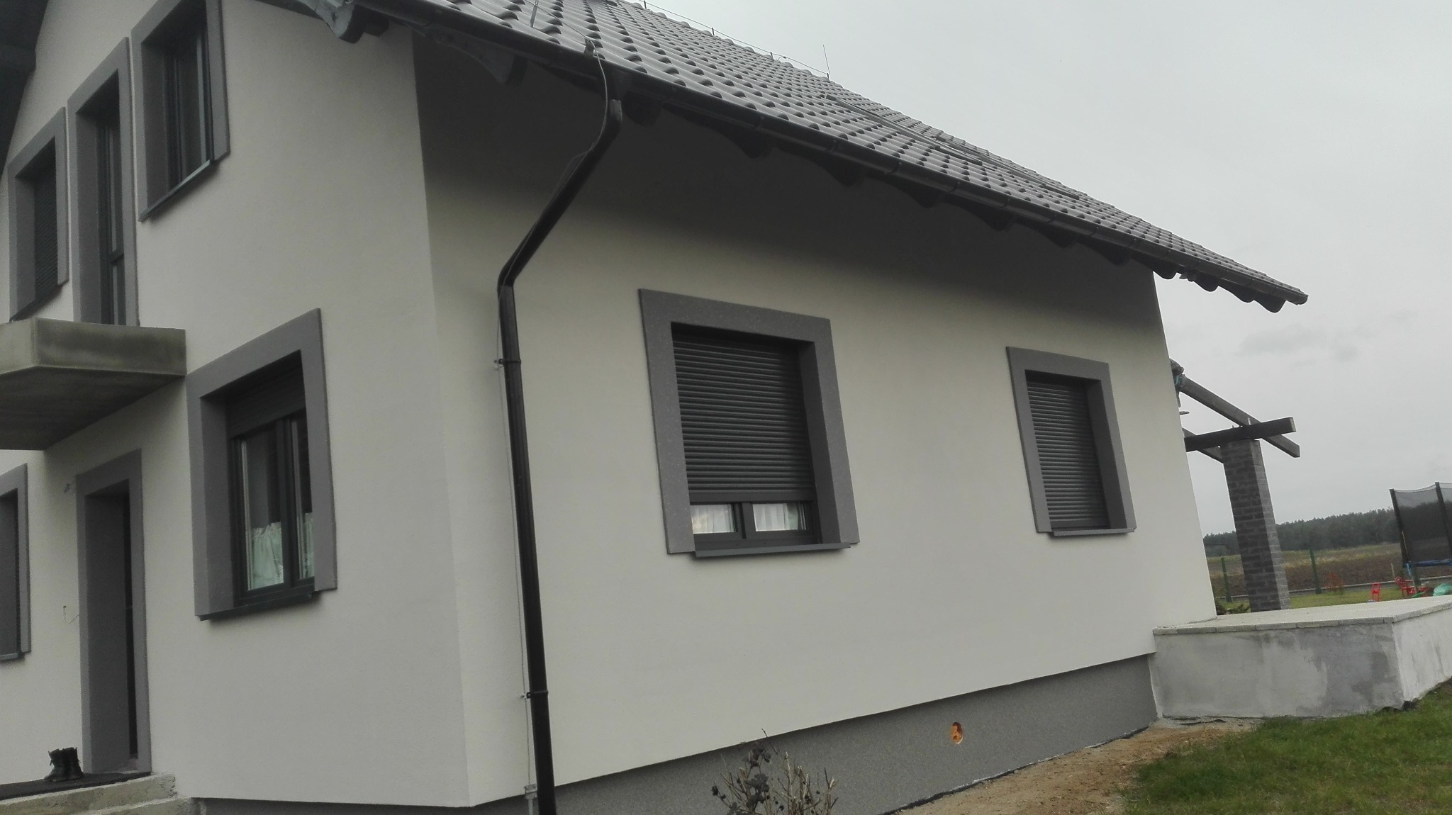 Elewacja domu jednorodzinnego w jasnych odcieniach szarości, z ciemnoszarą stolarką okienną i roletami zewnętrznymi, widoczny fragment dachu z ciemnej dachówki ceramicznej i rynna spustowa, w tle...