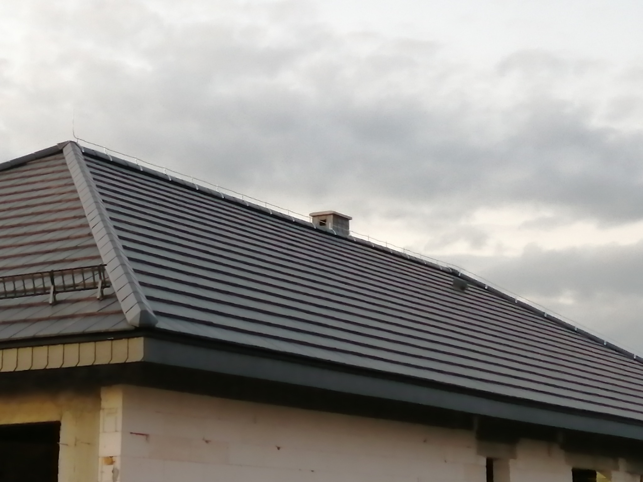 Nowy, grafitowy dach z dachówki ceramicznej na surowym budynku z białego pustaka, widoczny system rynnowy i komin.