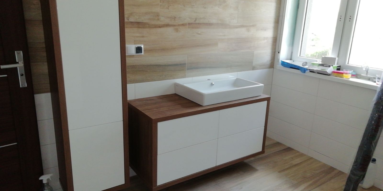 Nowoczesna łazienka z umywalką nablatową na szafce z białymi frontami i drewnianym blatem, drewnopodobne płytki na ścianie i podłodze, okno z akcesoriami malarskimi na parapecie.