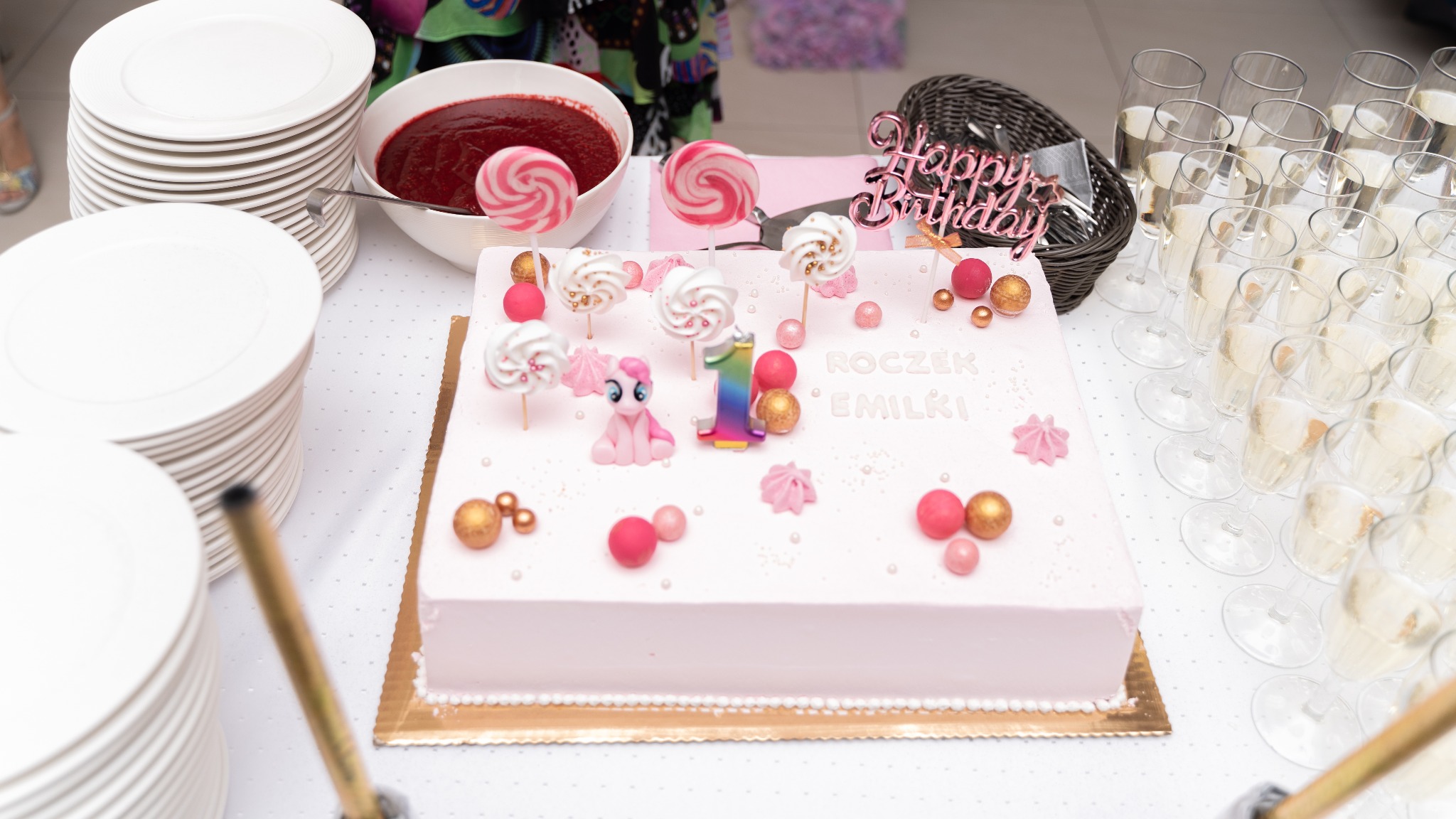 Tort urodzinowy dla dziecka z napisem 'Roczek Emilki', dekorowany lizakami, bezami, figurką konika Pony i świeczką w kształcie cyfry 1, obok stosy talerzy, miska z sosem i kieliszki z szampanem.