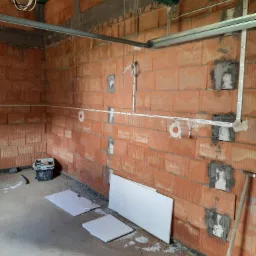 Surowa ściana z czerwonej cegły z widocznymi przewodami elektrycznymi w białych rurkach, metalowe profile sufitowe, fragment drewnianej konstrukcji stropu, wiadro z narzędziami i fragmenty płyt...