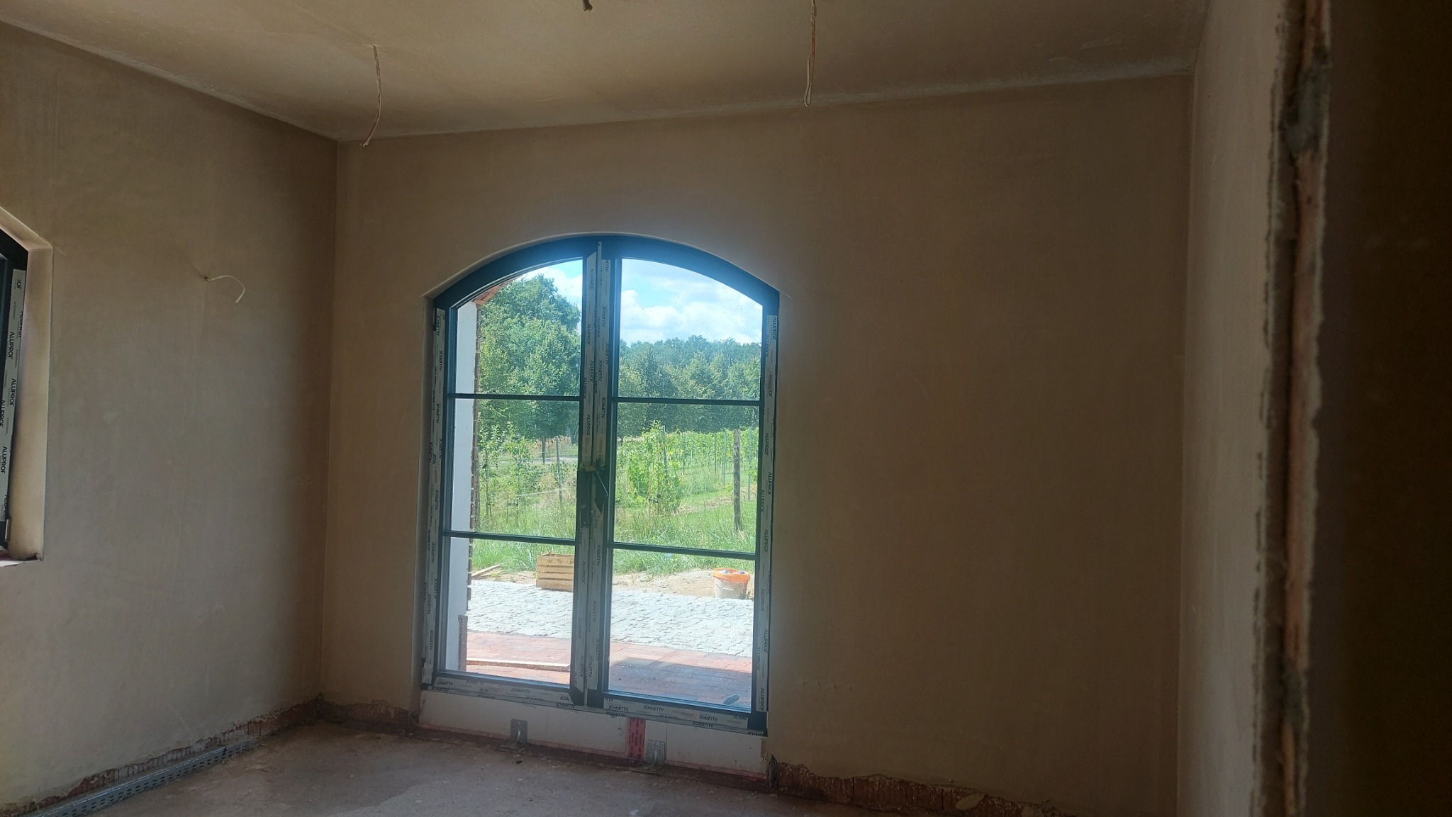 Wnętrze po nałożeniu tynku maszynowego, widoczne gładkie ściany i okno z widokiem na zieleń. Świeżo otynkowane ściany w jasnym odcieniu beżu.