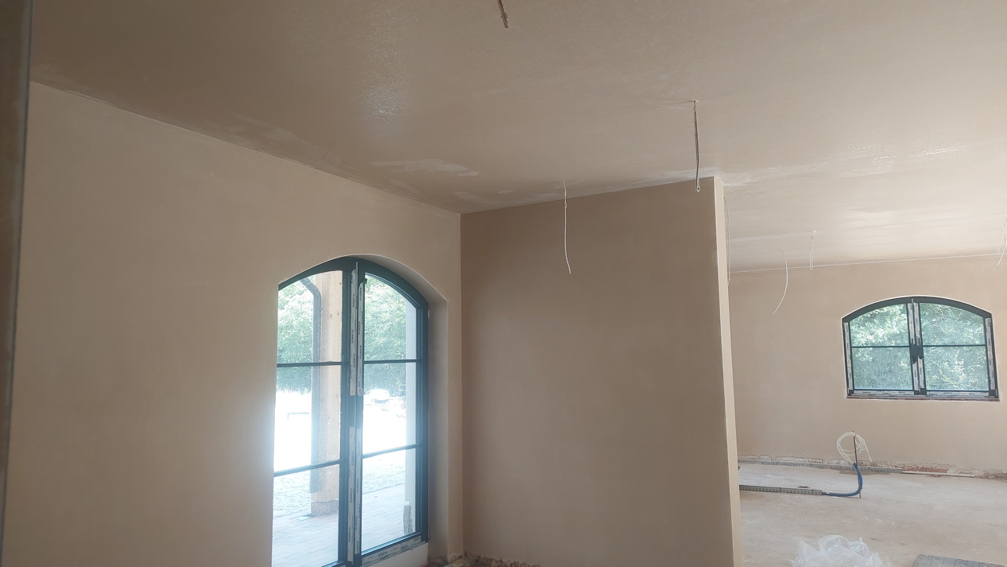 Wnętrze po nałożeniu gładzi gipsowej: ściany i sufit w jasnym odcieniu beżu, okna łukowe z widokiem na zieleń, wystające przewody elektryczne.