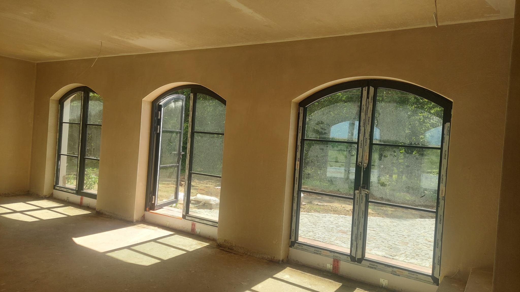 Wnętrze po nałożeniu tynków: trzy łukowe okna z czarnymi ramami, świeżo otynkowane ściany w odcieniach beżu, widoczne ślady prac budowlanych na podłodze.