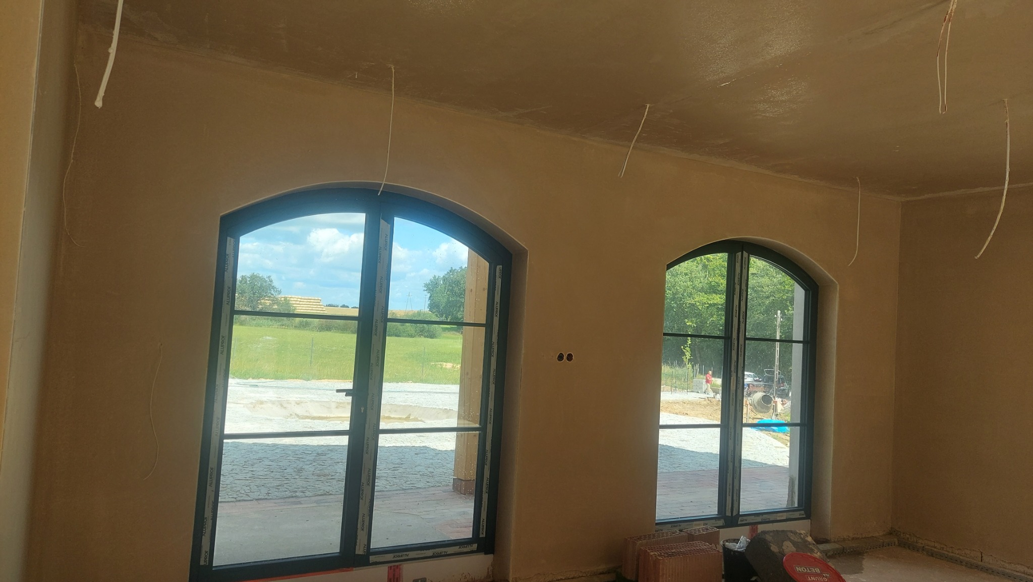 Wnętrze po tynkowaniu maszynowym: gładkie ściany w odcieniach beżu, dwa łukowe okna z widokiem na zewnątrz, wystające kable. Widoczne cegły i wiadro z napisem BETON.