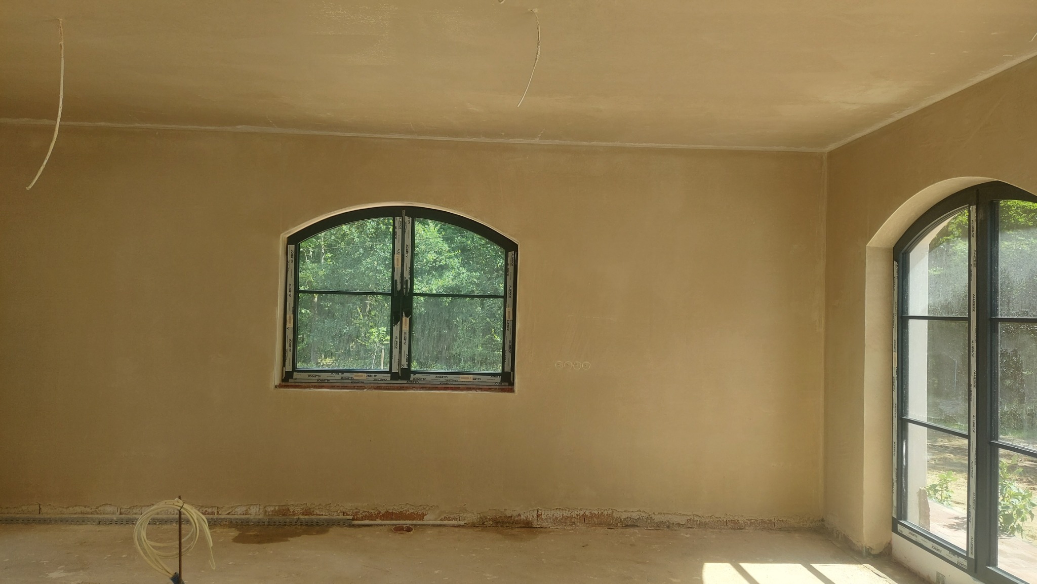 Wnętrze po nałożeniu tynków: gładkie, beżowe ściany z dwoma oknami łukowymi, przez które widać zieleń. Na podłodze widoczne ślady prac wykończeniowych.