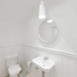 Jasna łazienka z białymi panelami ściennymi, okrągłym lustrem, umywalką na postumencie i toaletą, wyróżniająca się wzorzystą, patchworkową podłogą z płytek ceramicznych.