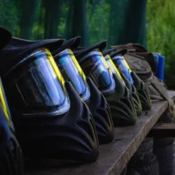 Rząd hełmów do paintballa, ustawionych na drewnianej ławce, z widocznymi odblaskami na wizjerach i leśnym tłem.