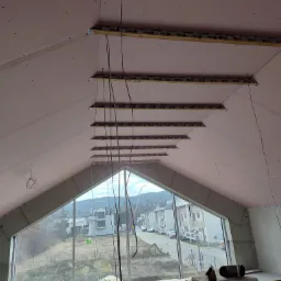 Poddasze z oknem szczytowym w trakcie montażu płyt karton-gips, widoczne profile montażowe i wystające przewody elektryczne, za oknem zabudowa jednorodzinna.