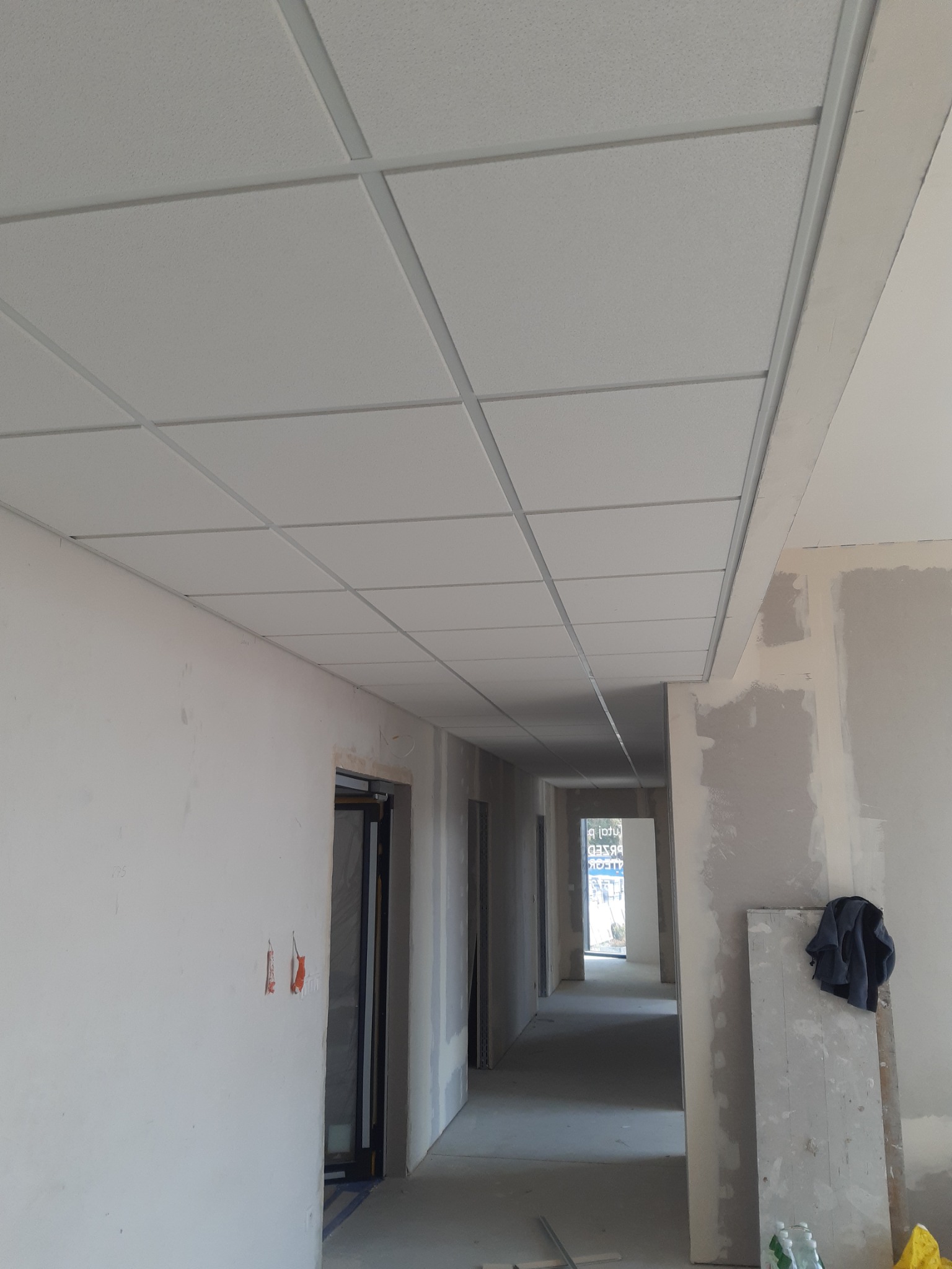 Sufit podwieszany kasetonowy z widoczną konstrukcją nośną w korytarzu budynku w stanie deweloperskim, ściany przygotowane do malowania.
