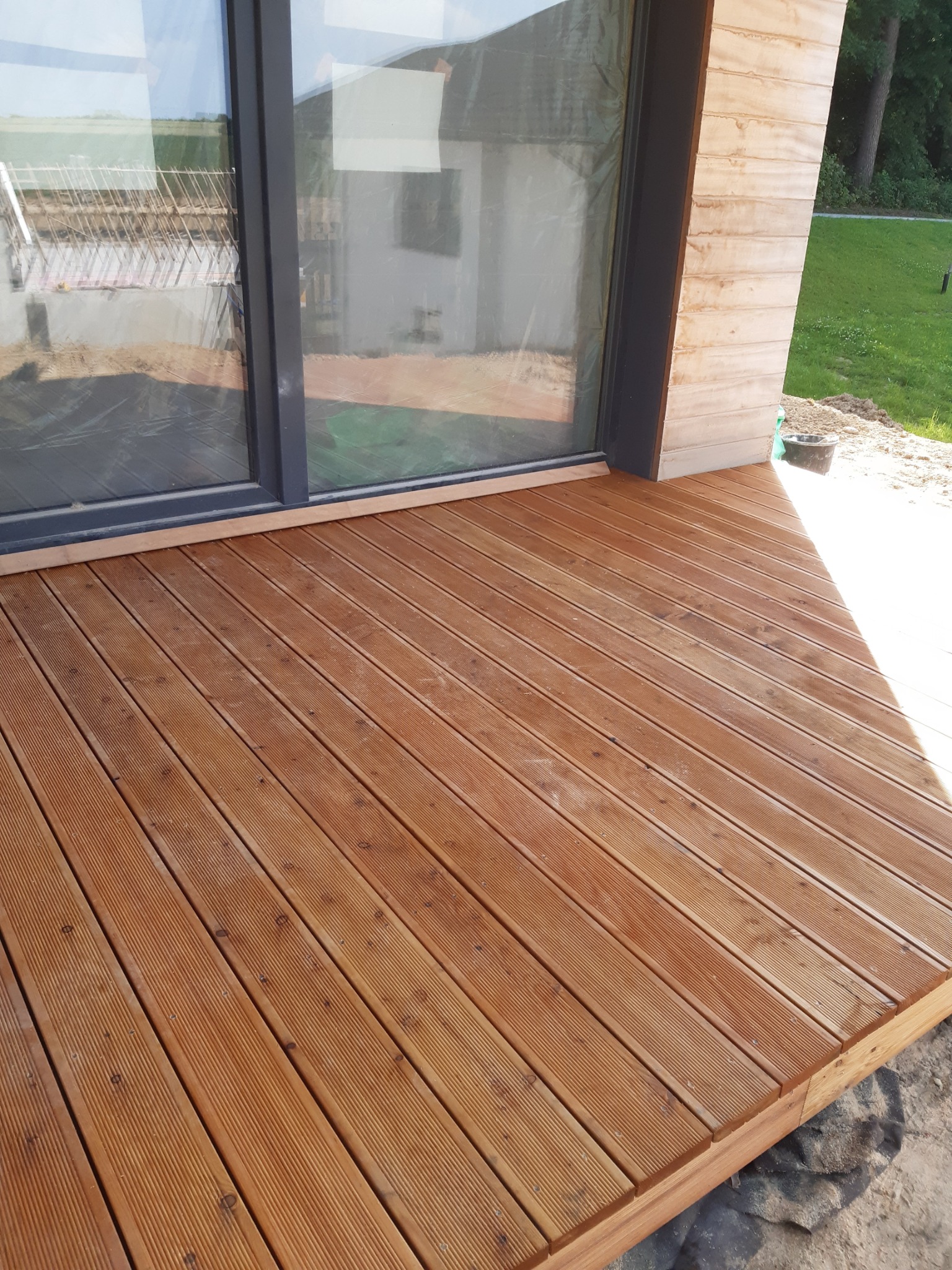 Wykończony taras z desek kompozytowych w kolorze ciepłego brązu, przylegający do nowoczesnego budynku z dużym oknem przesuwnym. Widoczne świeże nasadzenia trawy w tle.