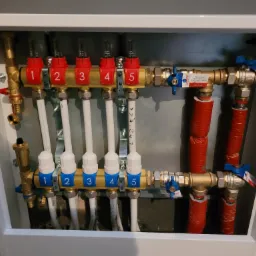 Rozdzielacz ogrzewania podłogowego w białej szafce, z pięcioma pętlami, zaworami termostatycznymi z numeracją 1-5, rurami PEX i zaworami kulowymi z niebieskimi motylkami.