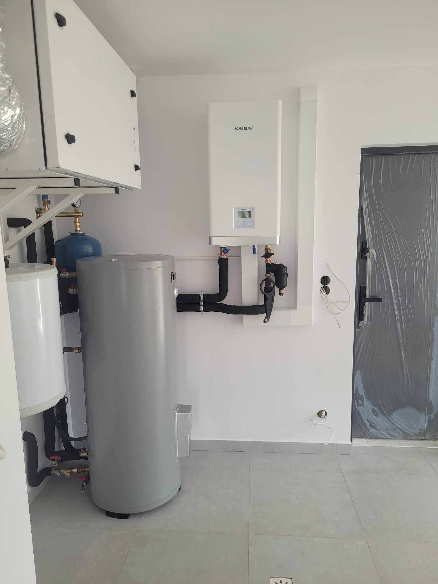 Biała kotłownia z piecem gazowym Kaisai, zasobnikiem ciepłej wody i instalacją hydrauliczną, widok na ścianę z przewodami elektrycznymi i drzwi zabezpieczone folią.
