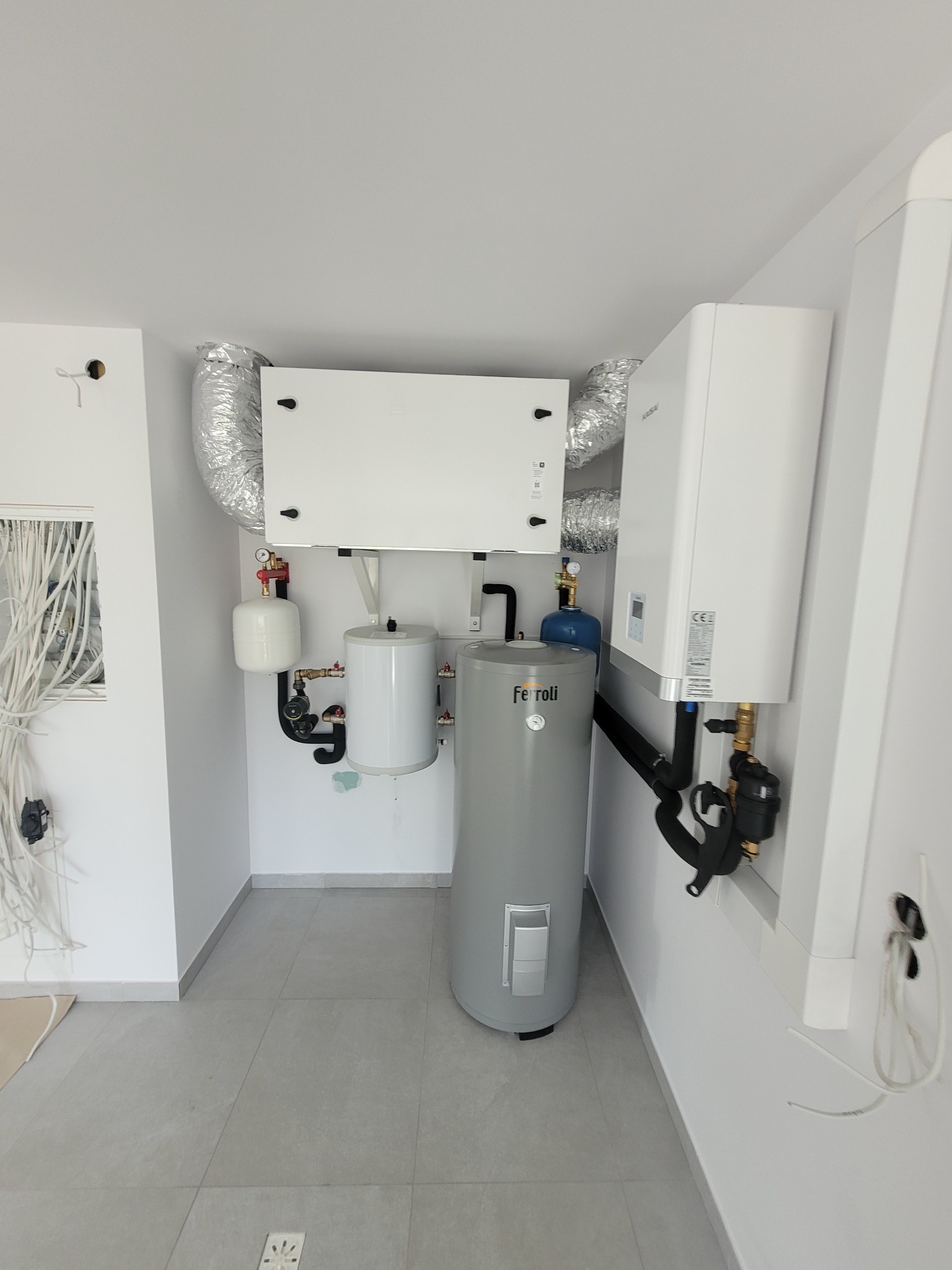 Wnętrze kotłowni z szarym zasobnikiem Ferroli, białym piecem i centralą wentylacyjną z aluminiowymi przewodami wentylacyjnymi, widoczne liczne rury i zawory.